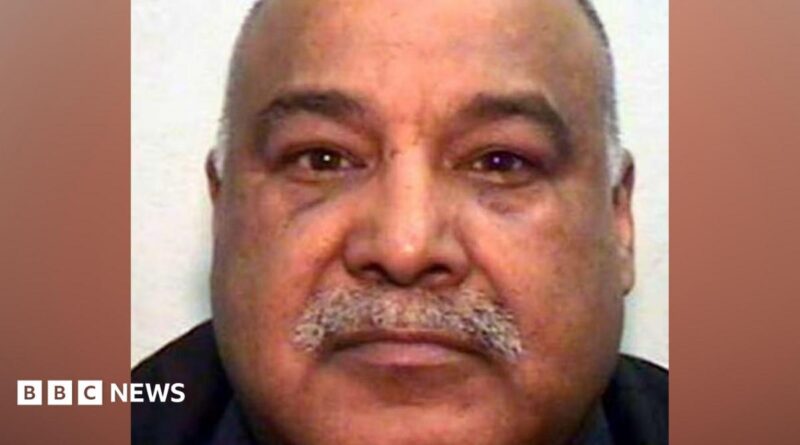A mugshot of an Asian man in his 60s, with a grey moustache and bald head. He is looking into the camera.