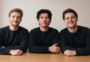 Three caucasian men in black long-sleeved crewneck shirts sit at a desk against a beige background.