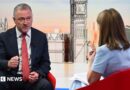 Housing, Communities and Local Government Secretary Steve Reed appearing on the Sunday with Laura Kuenssberg, with presenter Victoria Derbyshire standing in. The pair sit on red bucket chairs. Reed wears a black suit with a white shirt and a red tie. Derbyshire is wearing a pastel blue dress with short sleeves. She has shoulder length, dark blonde hair. 
