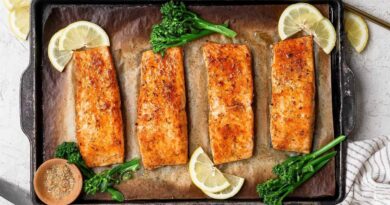 Broiled salmon fillets on a baking sheet garnished with lemon slices, broccolini, and a bowl of seasoning.