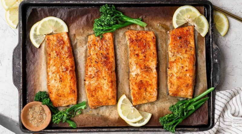 Broiled salmon fillets on a baking sheet garnished with lemon slices, broccolini, and a bowl of seasoning.
