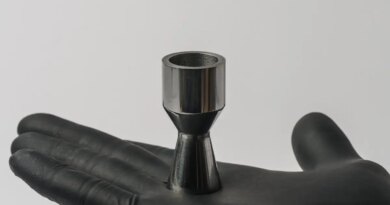 A gloved hand holds a metal part shaped like a rocket nozzle.