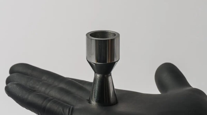 A gloved hand holds a metal part shaped like a rocket nozzle.