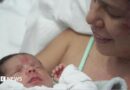 Dayana Patino grins as she holds her son Juan David close to her chest from a hospital bed. Her husband, Gerson, stands beside them wearing a light blue t-shirt. 