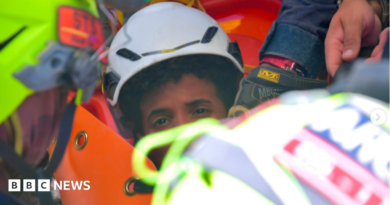 the picture shows half of the boys face - the rest is obscured by rescue workers. He's wearing a white hard hat and has dark curly hair.