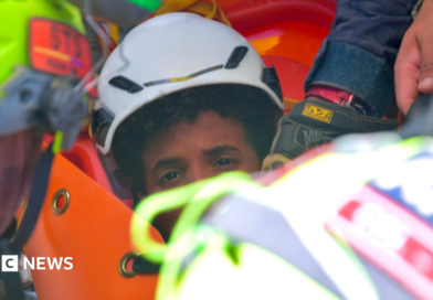 the picture shows half of the boys face - the rest is obscured by rescue workers. He's wearing a white hard hat and has dark curly hair.