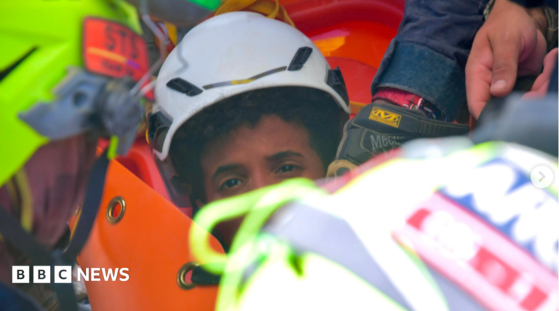 the picture shows half of the boys face - the rest is obscured by rescue workers. He's wearing a white hard hat and has dark curly hair.