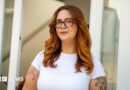 Jess King, a woman with long ginger hair and glasses, stands in front of a doorway. She is wearing a short-sleeved white top and has visible black tattoos at the top of her arms. 
