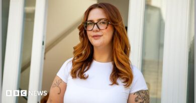 Jess King, a woman with long ginger hair and glasses, stands in front of a doorway. She is wearing a short-sleeved white top and has visible black tattoos at the top of her arms.
