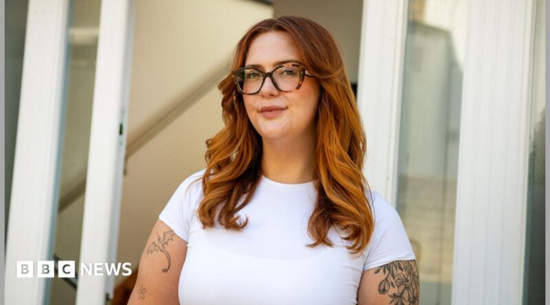 Jess King, a woman with long ginger hair and glasses, stands in front of a doorway. She is wearing a short-sleeved white top and has visible black tattoos at the top of her arms.