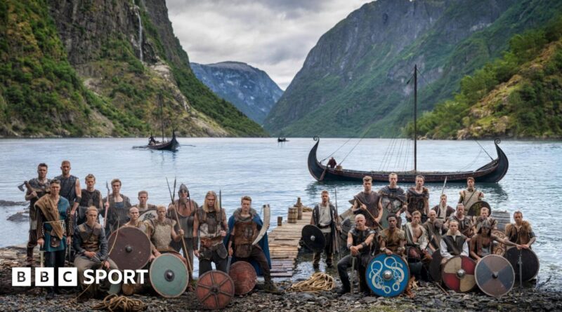 Norway players in Viking dress and with weapons and shields pose in front of a fjord with Viking boats