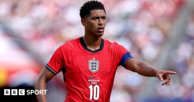 Jude Bellingham, wearing a red England shirt with a white number 10 in the centre beneath the badge, extends his left arm and points his index finger