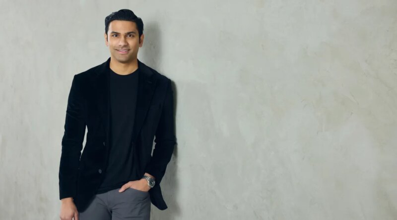 A well-dressed man leans against a neutral colored wall