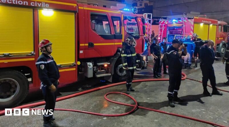 Firefighters in blue uniforms stand in front of fire trucks