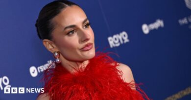 A zoomed in, slanted angle of Amber Davies, in a red feathered dress and wearing big pearl earrings. She stands in front of a branded blue wall with sponsor logos. Her dark hair is pulled back into a slick bun.