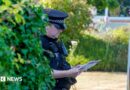A police officer looks down at a sheet of paper while wearing uniform next to a bush and police van outside Widdecombe's home on Friday.