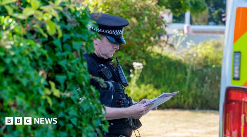 A police officer looks down at a sheet of paper while wearing uniform next to a bush and police van outside Widdecombe's home on Friday.