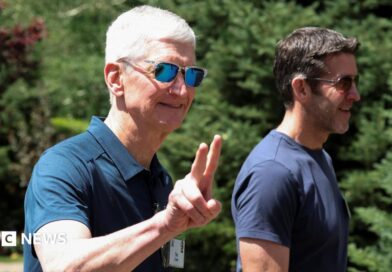 Apple CEO Tim Cook walking outside, throwing up 'peace' fingers, wearing a blue polo shirt and reflective sunglasses. John Ternus, who will replace Cook as CEO later this year, walks beside him, wearing a blue t-shirt and sunglasses.