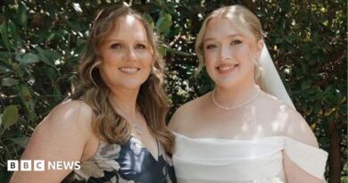 Hannah and her mother at Hannah's wedding. Her mother is wearing a blue dress with white flowers. Hannah on the right is dressed in a white wedding dress holding a bouquet