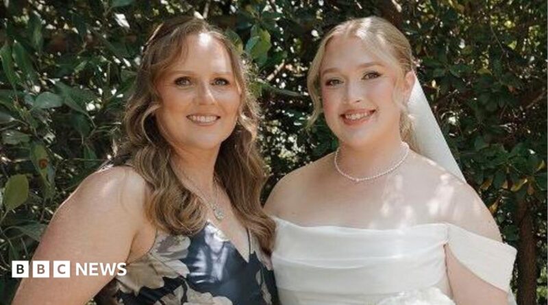 Hannah and her mother at Hannah's wedding. Her mother is wearing a blue dress with white flowers. Hannah on the right is dressed in a white wedding dress holding a bouquet