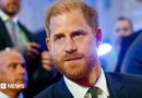 Headshot of Prince Harry in a blue suit, white shirt, and dark green tie looking off-camera in a room with several men in suits out of focus in the background,