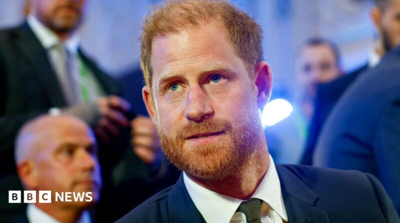Headshot of Prince Harry in a blue suit, white shirt, and dark green tie looking off-camera in a room with several men in suits out of focus in the background,
