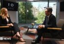 Andy Burnham, wearing a dark suit, gestures with his hands while speaking during an interview. Behind him is a TV with the logo for No 10 North.