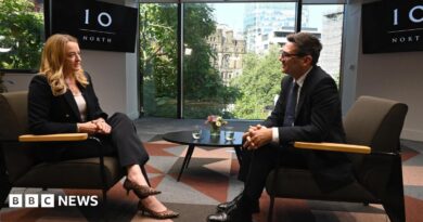 Andy Burnham, wearing a dark suit, gestures with his hands while speaking during an interview. Behind him is a TV with the logo for No 10 North.
