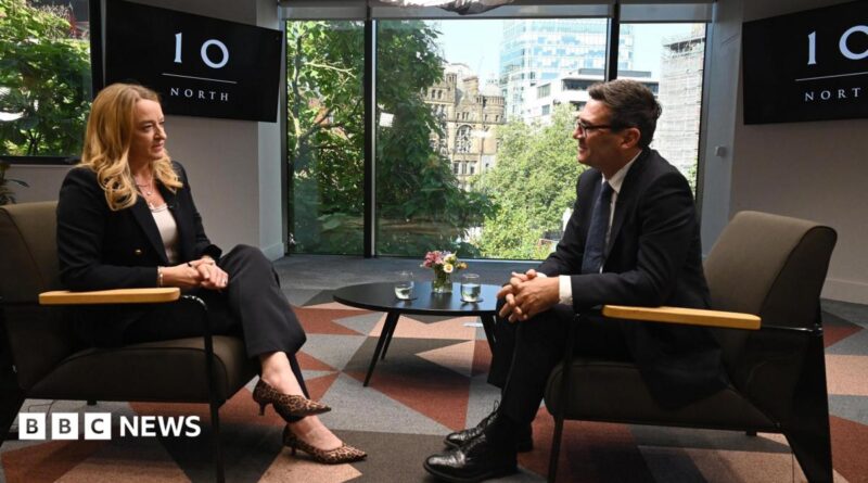 Andy Burnham, wearing a dark suit, gestures with his hands while speaking during an interview. Behind him is a TV with the logo for No 10 North.
