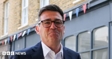 Andy Burnham looks directly forward and smiles with his mouth closed. He wears tortoise shell glasses, a navy suit jacket is navy and a white shirt. He stands in a street, behind him is a blue shop, with red, blue and white bunting hung above it.