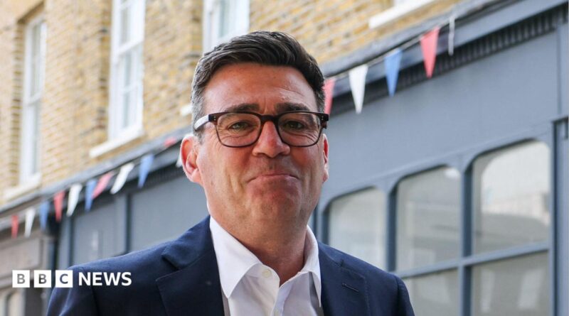 Andy Burnham looks directly forward and smiles with his mouth closed. He wears tortoise shell glasses, a navy suit jacket is navy and a white shirt. He stands in a street, behind him is a blue shop, with red, blue and white bunting hung above it.