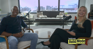 TechCrunch Build Mode podcast host Isabelle Johannessen sits across from venture capitalist Charles Hudson in a bright podcast studio. Both are smiling toward the camera, seated in armchairs with microphones positioned between them. A coffee table with two mugs sits in the foreground, while large windows behind them overlook the San Francisco skyline. The TechCrunch Build Mode logo appears in the bottom-right corner.