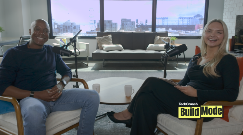 TechCrunch Build Mode podcast host Isabelle Johannessen sits across from venture capitalist Charles Hudson in a bright podcast studio. Both are smiling toward the camera, seated in armchairs with microphones positioned between them. A coffee table with two mugs sits in the foreground, while large windows behind them overlook the San Francisco skyline. The TechCrunch Build Mode logo appears in the bottom-right corner.
