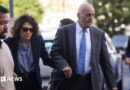 A balding man in a grey suit holds the hand of a woman in a navy blue suit who is wearing sunglasses as both are walking