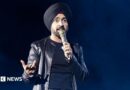Diljit Dosanjh kicks off his Aura World Tour at BC Place on April 23, 2026, in Vancouver, British Columbia, Canada. He is wearing a black jacket over a black t-shirt and sporting a black turban, as he speaks on a microphone from the stage.