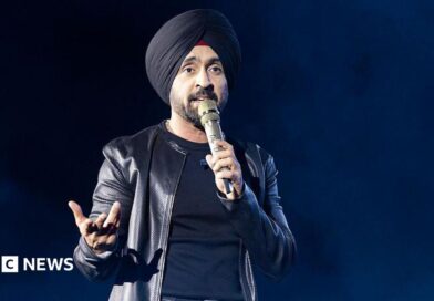 Diljit Dosanjh kicks off his Aura World Tour at BC Place on April 23, 2026, in Vancouver, British Columbia, Canada. He is wearing a black jacket over a black t-shirt and sporting a black turban, as he speaks on a microphone from the stage.