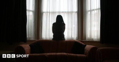A woman sits on the back of a sofa, looking out of the window in fear of her partner, coming home after football.