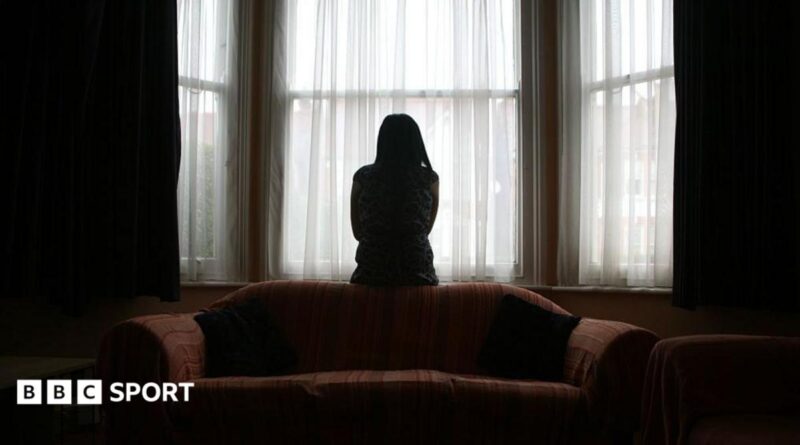 A woman sits on the back of a sofa, looking out of the window in fear of her partner, coming home after football.