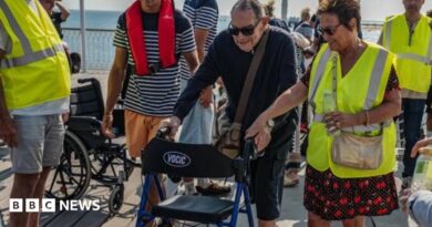 A man using a mobility aid walker is helped by volunteers dressed in yellow hi-vis jackets with the sea visible in the background.