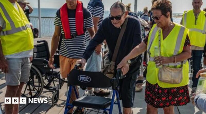A man using a mobility aid walker is helped by volunteers dressed in yellow hi-vis jackets with the sea visible in the background.