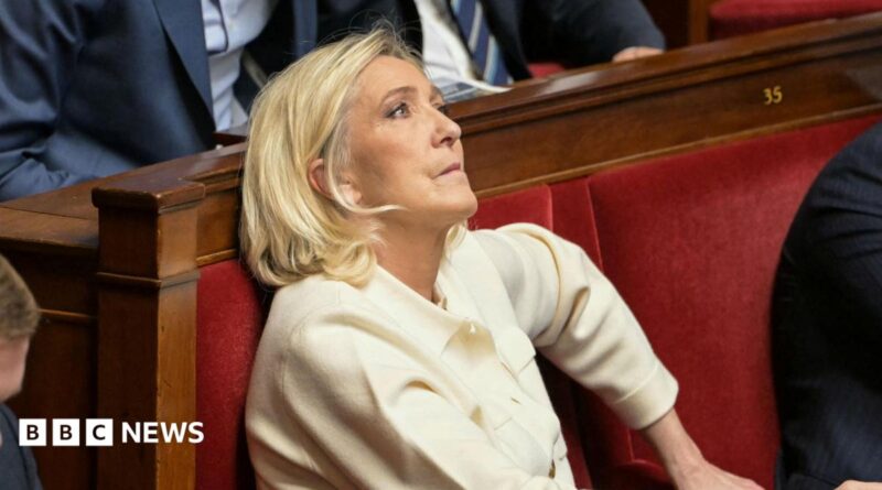 Marine Le Pen sits in France's National Assembly in a red velvet seat. She is biting her lip and looking up. She wears a cream suit.