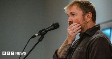 Graham Platner seen on stage at a microphone. his hand is over his mouth 