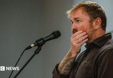 Graham Platner seen on stage at a microphone. his hand is over his mouth 
