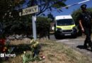 A police officer is seen walking beneath a cordon with a police van in the background and to the left a Drive Slowly sign with flowers underneath it.