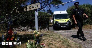 A police officer is seen walking beneath a cordon with a police van in the background and to the left a Drive Slowly sign with flowers underneath it.