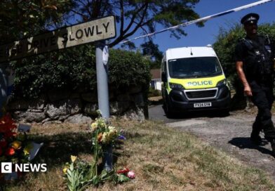 A police officer is seen walking beneath a cordon with a police van in the background and to the left a Drive Slowly sign with flowers underneath it.