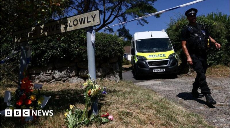 A police officer is seen walking beneath a cordon with a police van in the background and to the left a Drive Slowly sign with flowers underneath it.