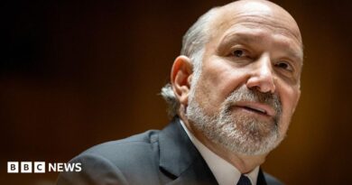 Close-up portrait of Howard Lutnick with a short grey beard and receding hairline, wearing a dark suit jacket, white shirt, and dark tie. The person is turned slightly to the side and looking toward the camera. The background is softly blurred with warm brown and amber tones, creating a shallow depth-of-field effect.