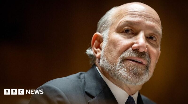 Close-up portrait of Howard Lutnick with a short grey beard and receding hairline, wearing a dark suit jacket, white shirt, and dark tie. The person is turned slightly to the side and looking toward the camera. The background is softly blurred with warm brown and amber tones, creating a shallow depth-of-field effect.