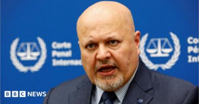 International Criminal Court Prosecutor Karim Khan speaks during an interview while sitting in front of a blue sign with blurry white writing and the symbol of the ICC - the scales of justice surrounded by a laurel wreath. 